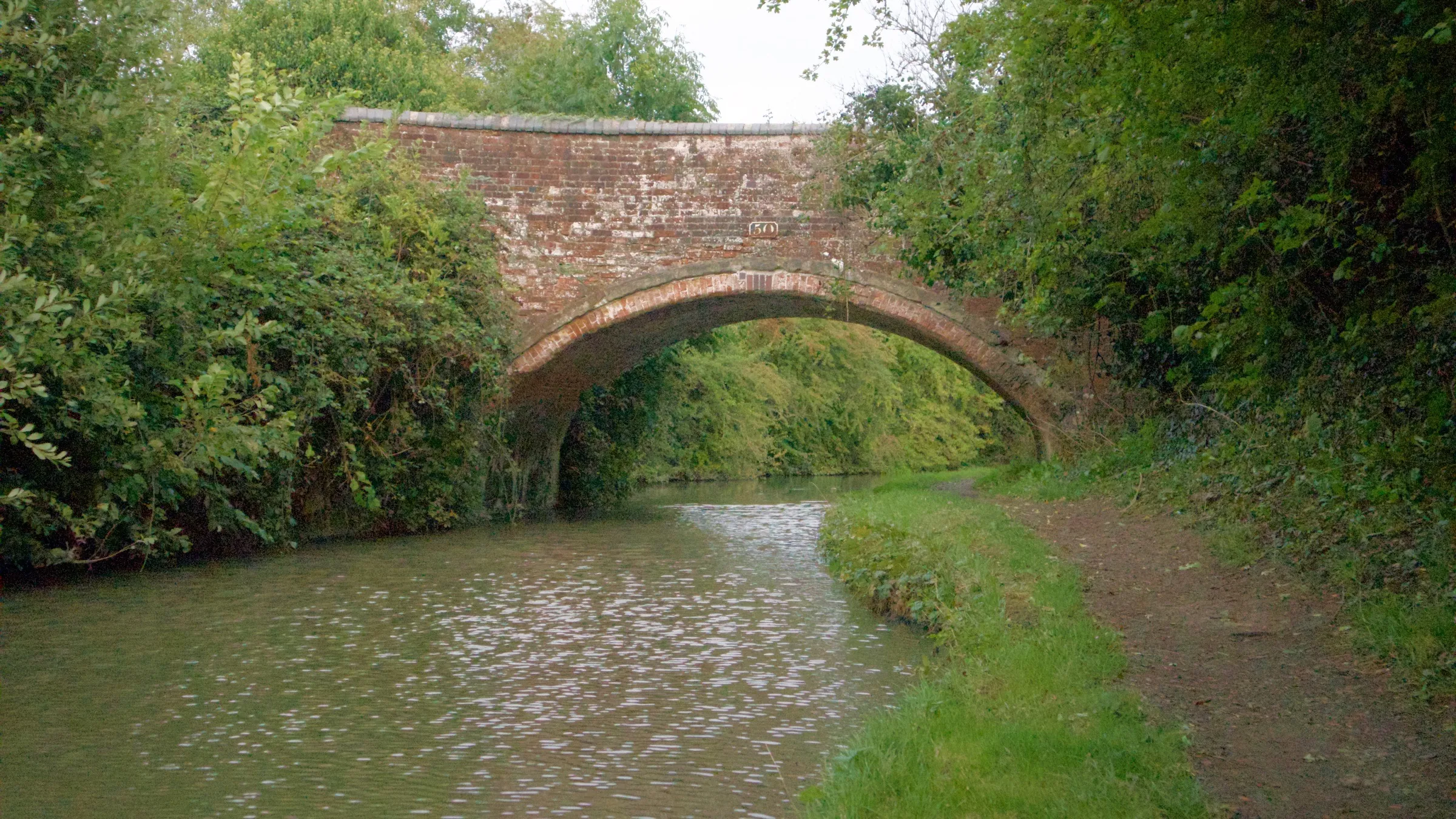 A brick bridge over a canal, with a towpath curving beneath the arch and trees on both banks