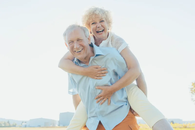 An older couple smiling outdoors, with the woman on the man’s back
