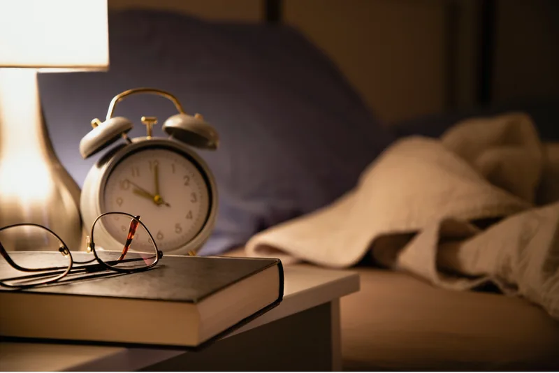 An alarm clock, glasses and a book on a bedside table beside a bed
