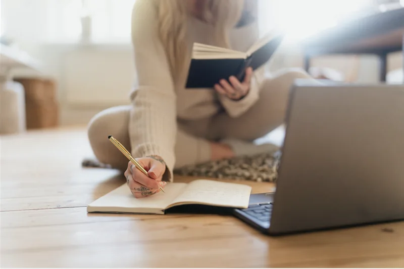 A person reading a book and making notes beside a laptop